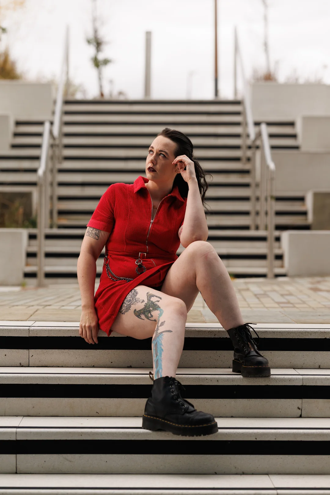 A person with tattoos in a red corduroy dress and black boots sitting on outdoor concrete stairs.