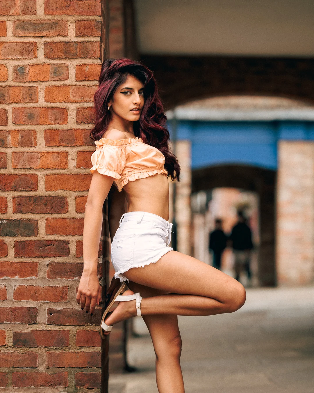 Woman with burgundy hair posing against a brick wall in a peach crop top and white denim shorts.