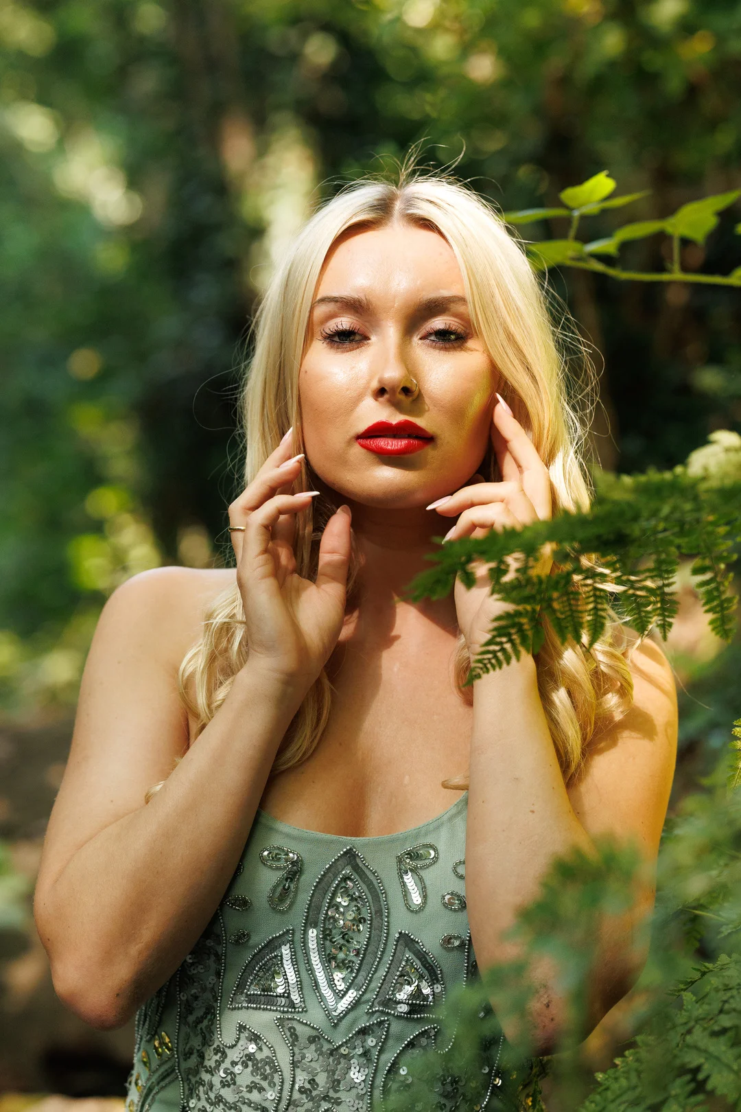 Blonde woman with red lipstick in a sequined dress posing in a lush green forest.