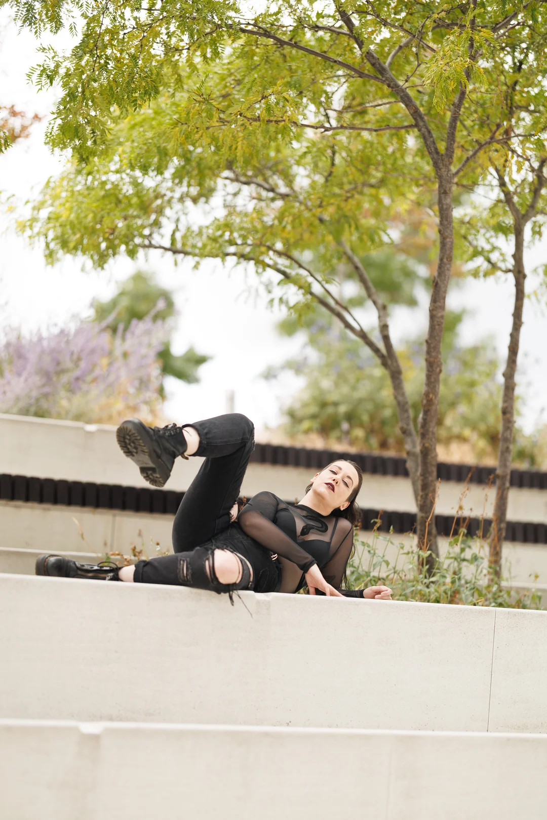 Woman in a black sheer top and ripped jeans posing on concrete steps with green trees behind her.
