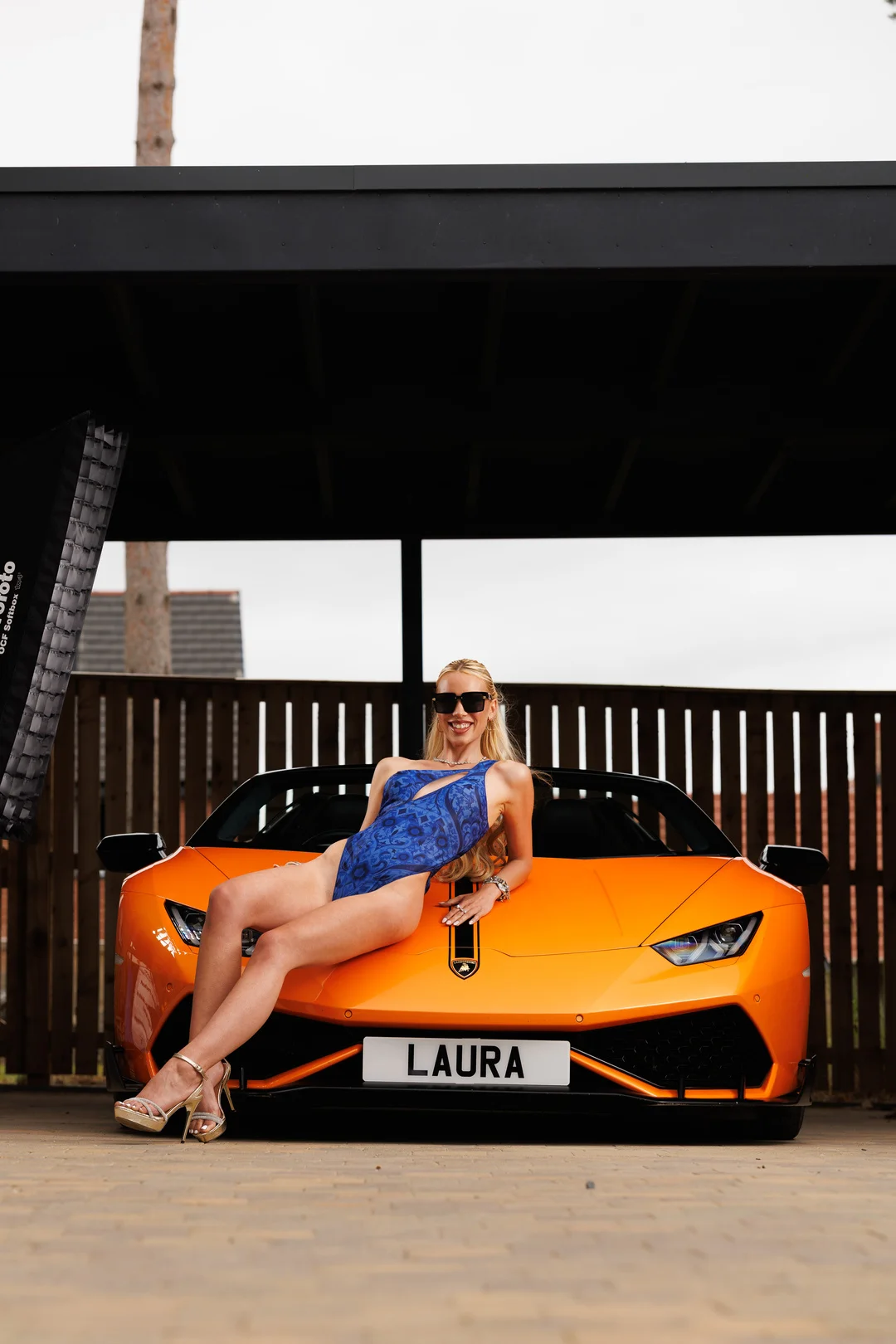 A woman in a blue swimsuit posing on the hood of an orange Lamborghini under a carport.