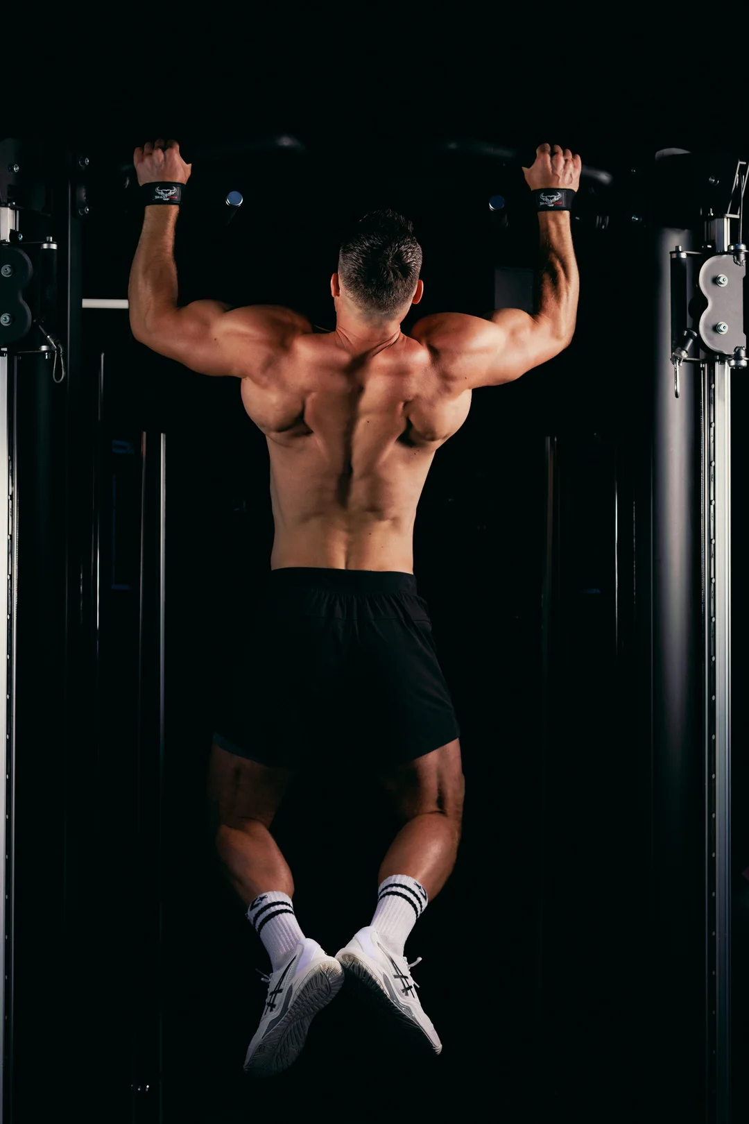 Rear view of a muscular man performing a pull-up in a gym with dramatic dark lighting.