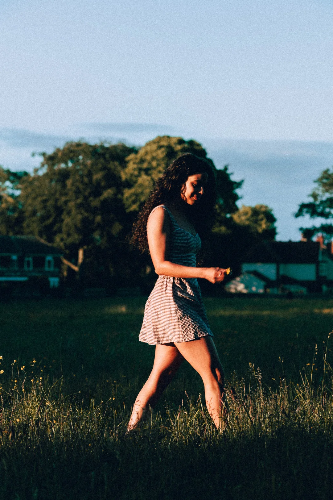 A woman with curly hair in a short checkered dress walking through a grassy field at golden hour.