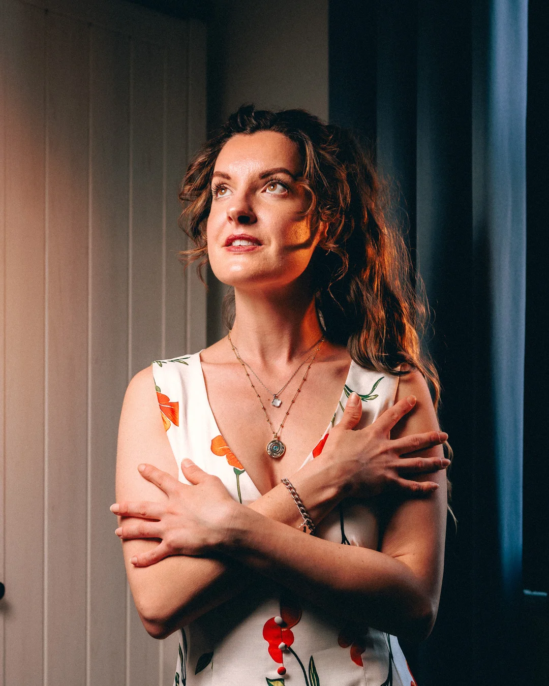 A pensive woman in a white floral dress looking upwards with her arms crossed over her chest.
