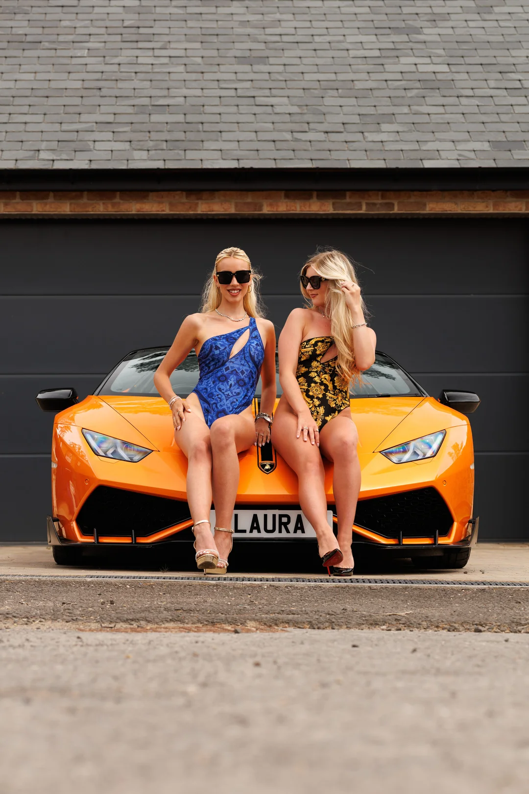 Two blonde women in patterned swimsuits posing on the hood of an orange luxury sports car.
