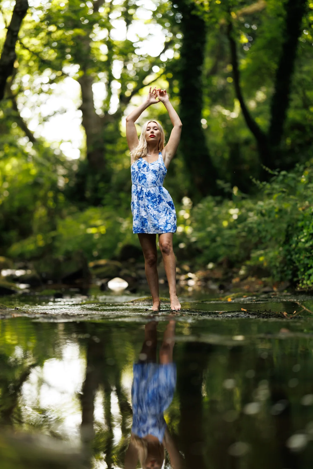 Woman in a blue floral dress posing in a forest stream with a reflection in the water.
