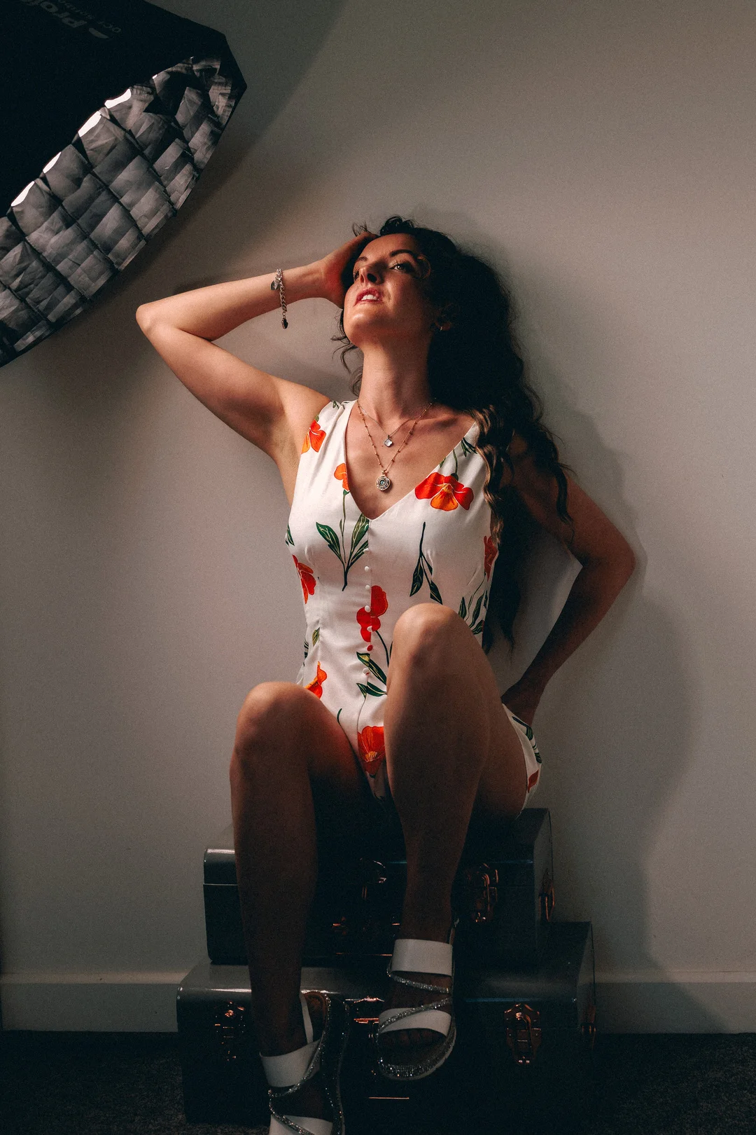 Woman in a white floral dress posing on vintage suitcases with studio lighting visible.