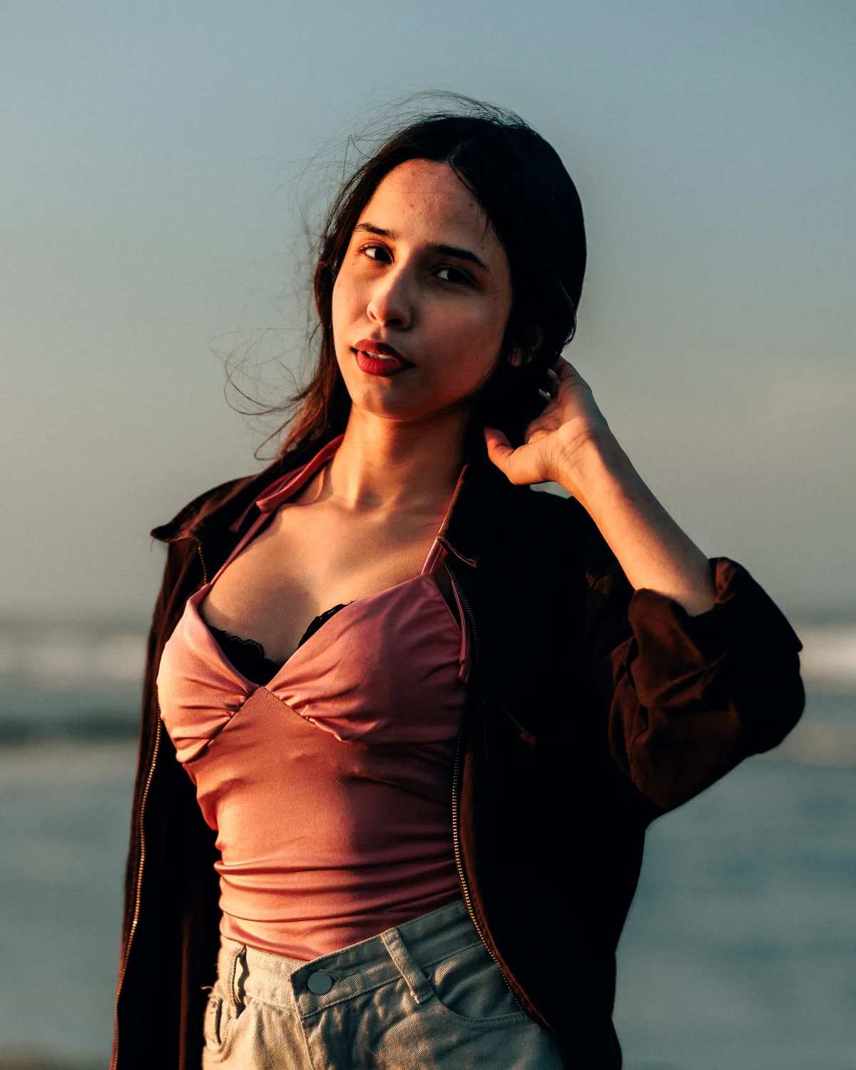 Portrait of a young woman in a pink satin top and dark jacket posing outdoors during golden hour.