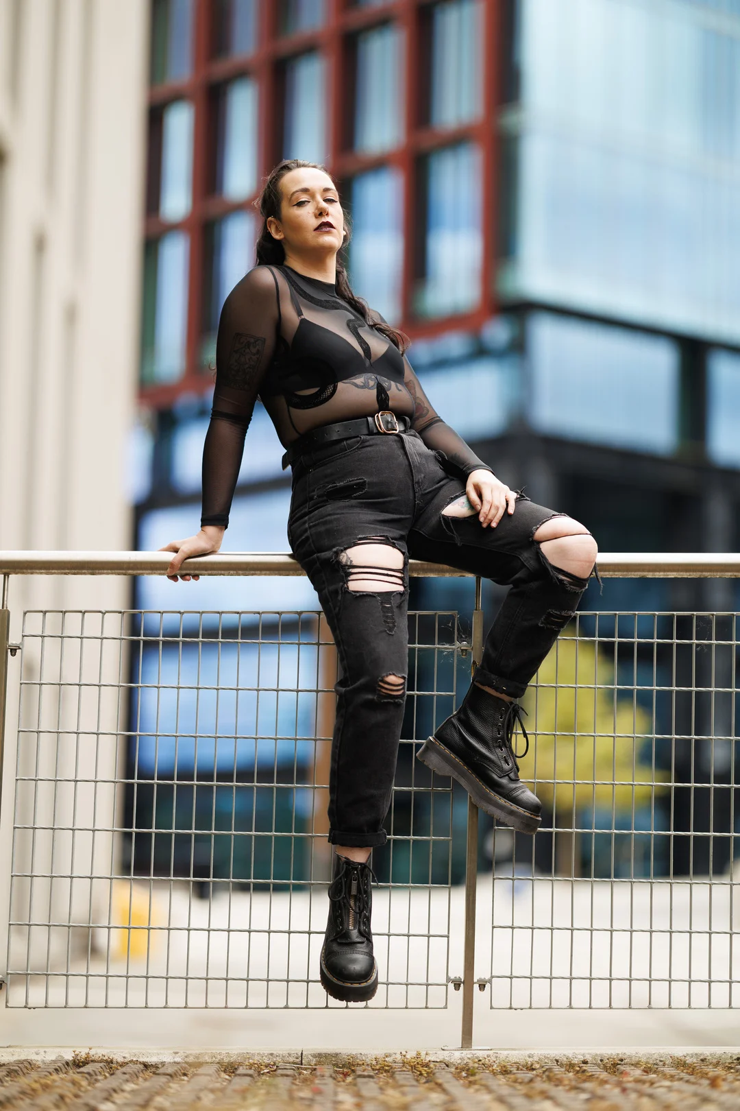 A woman in a black sheer top and ripped black jeans posing against a metal railing in an urban setting.