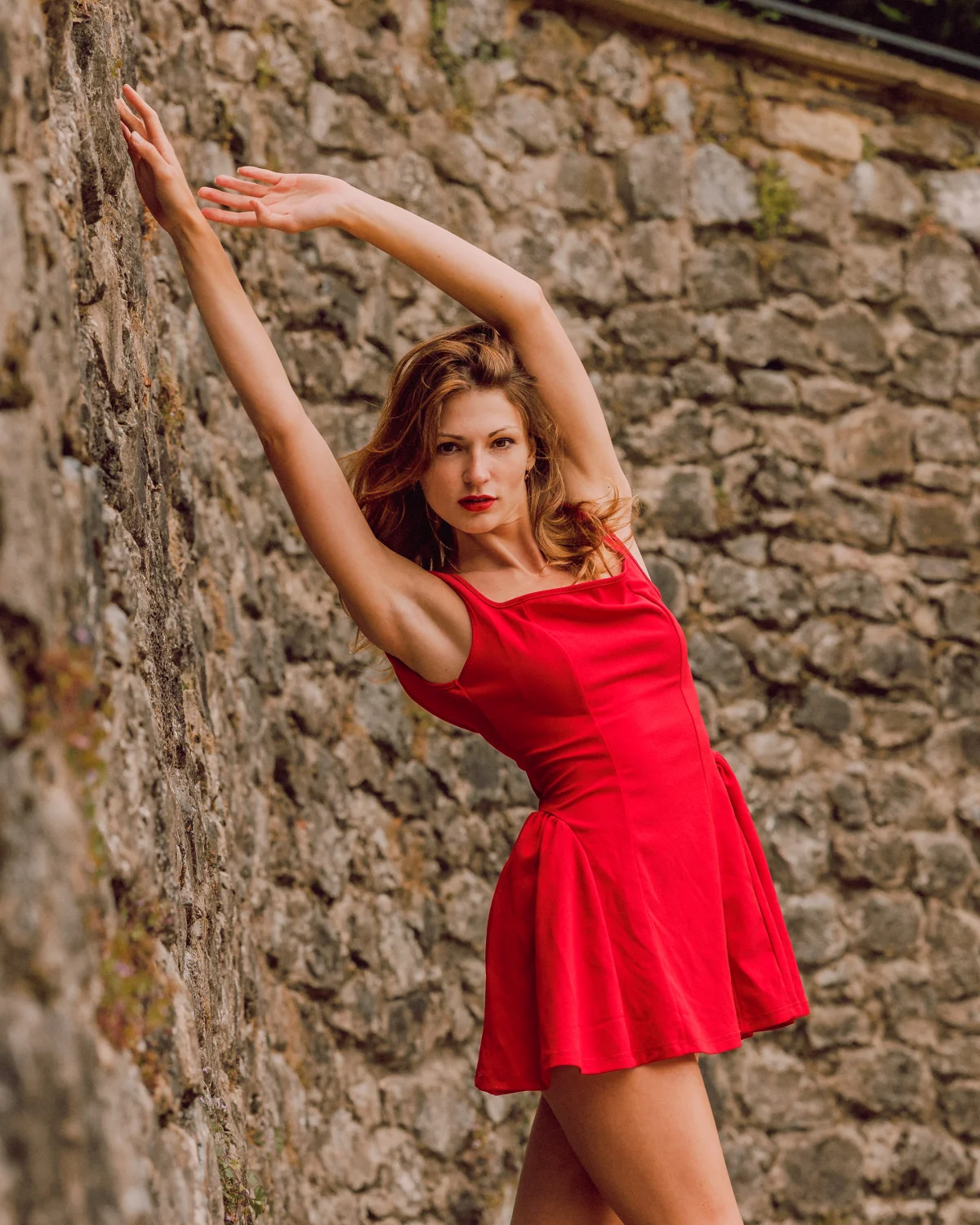 A woman in a short red dress posing against a rustic stone wall, looking confidently at the camera.
