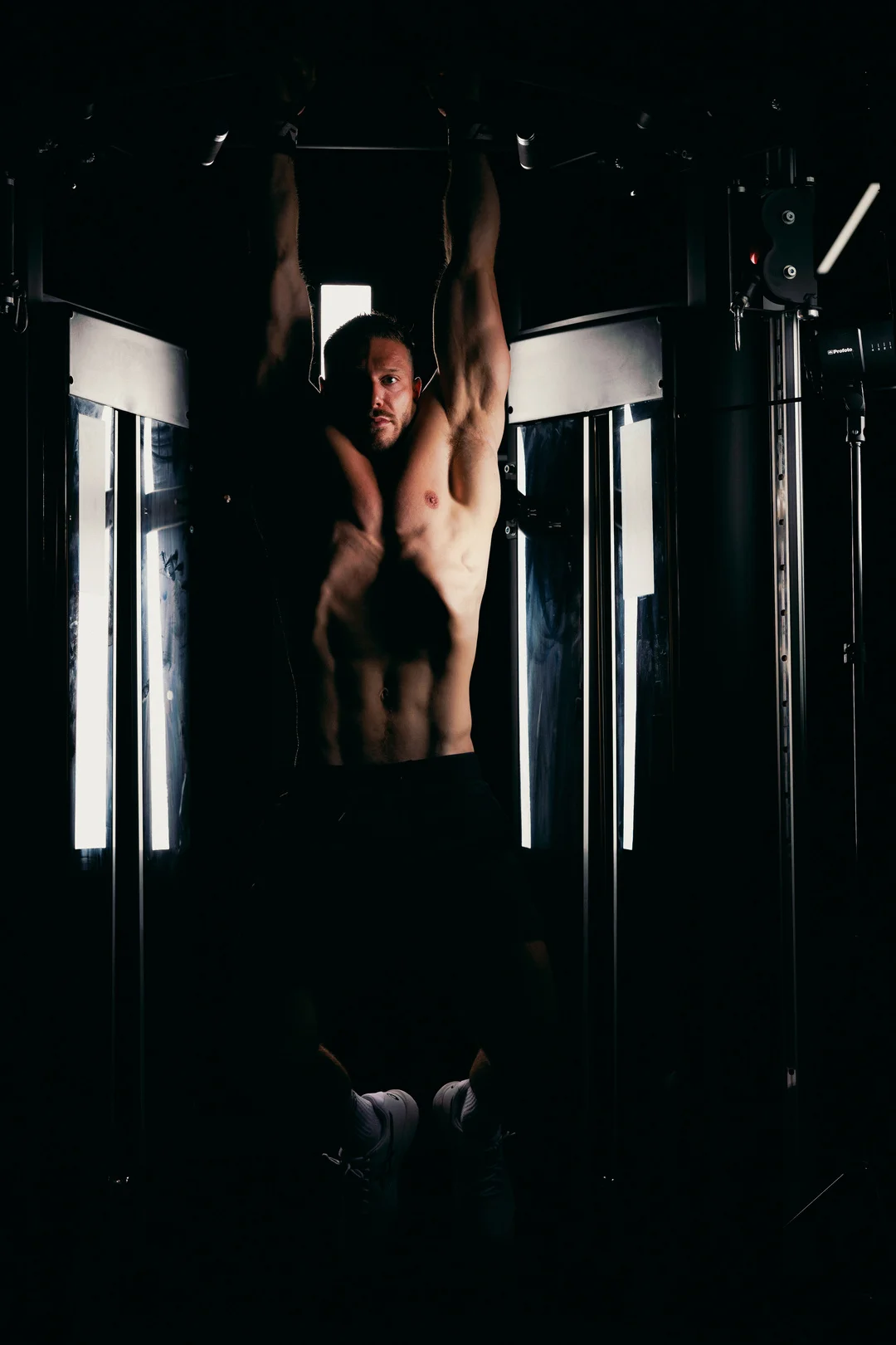 A muscular man hanging from a pull-up bar in a dark gym with dramatic side lighting.