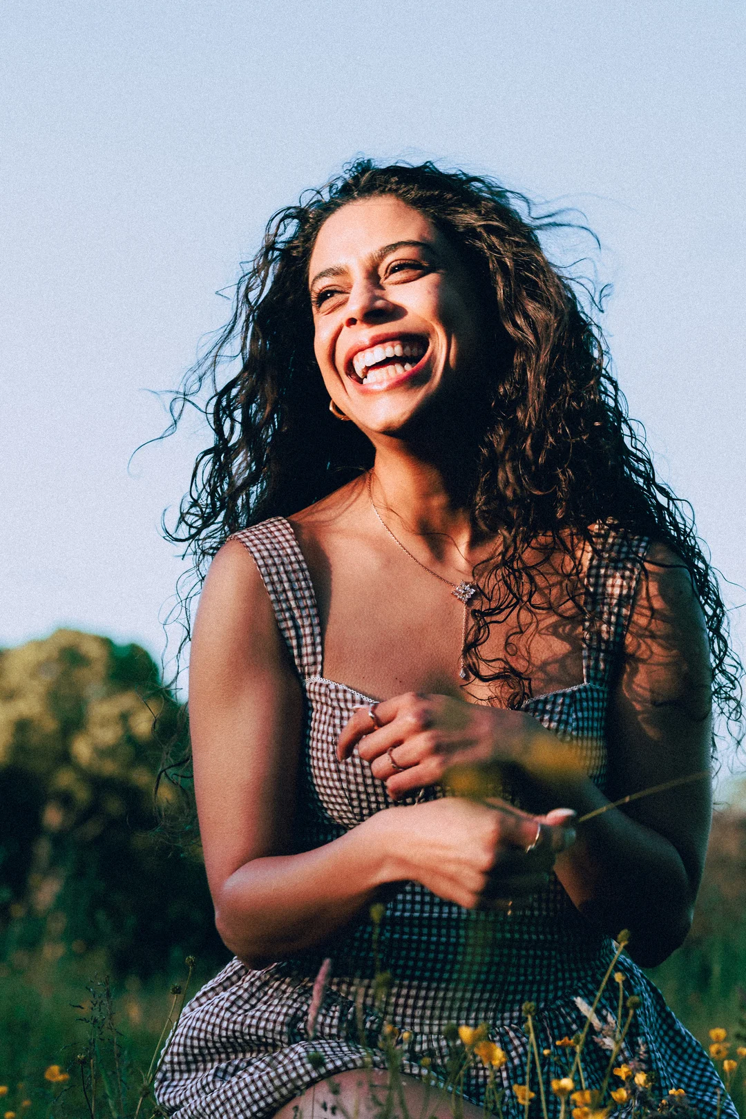 A laughing woman with curly hair wearing a gingham dress in a sunny outdoor field.