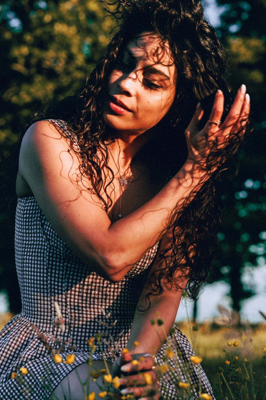 A woman with long curly hair in a gingham dress posing in a sun-drenched field during golden hour.