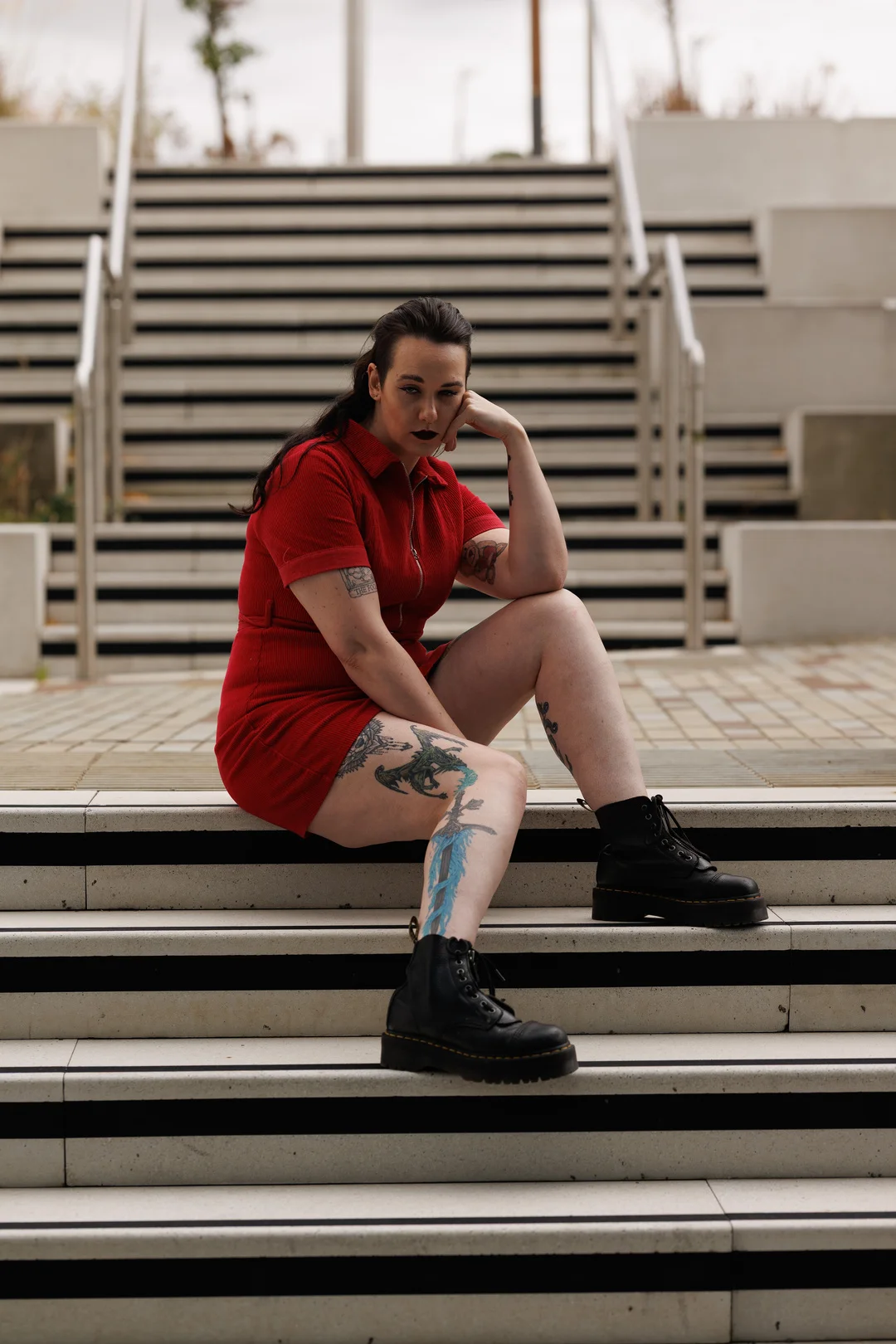 A woman with dark hair and tattoos wearing a red dress and black boots sitting on outdoor stairs.