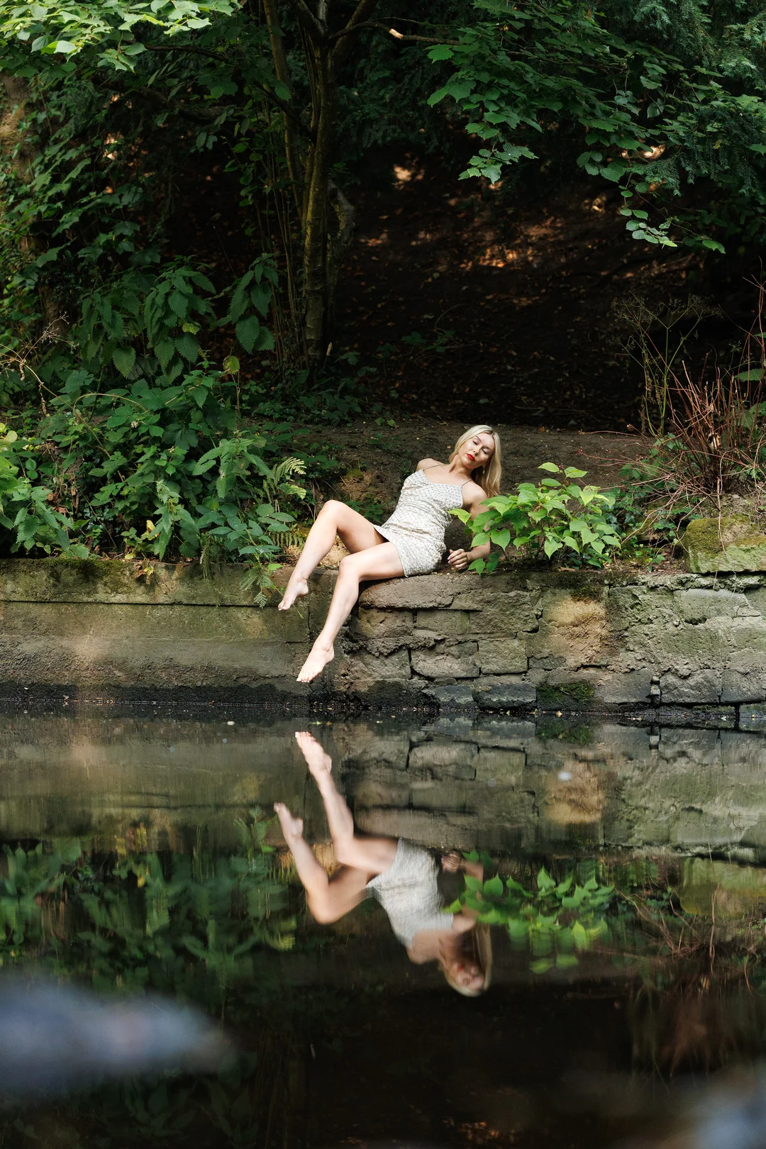 Blonde woman in a short white dress posing on a stone ledge by a pond, with a clear reflection in the water.