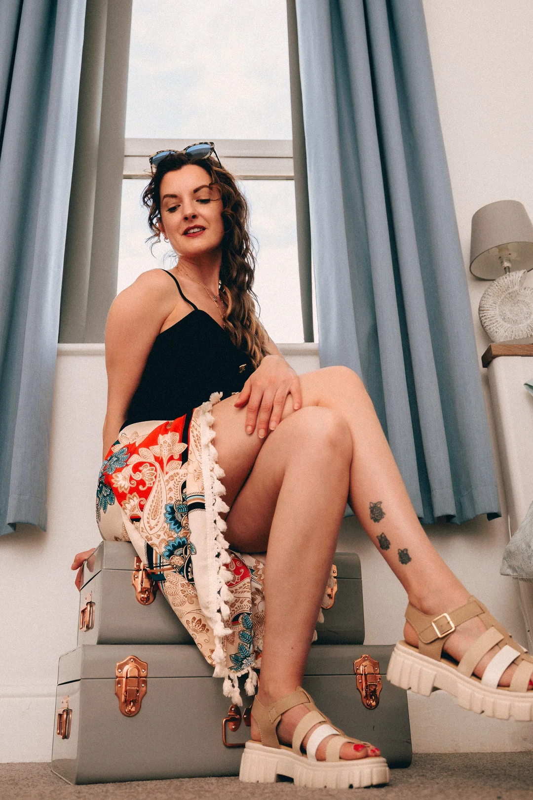 Woman in a black top and colorful patterned skirt posing on grey vintage suitcases by a window.