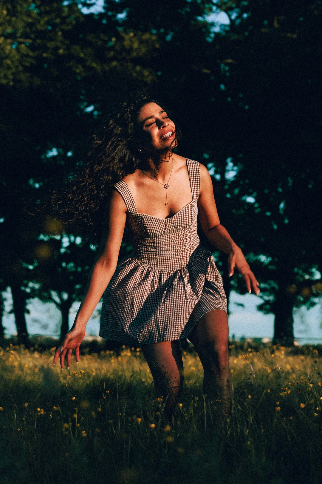 A woman with curly hair in a gingham dress laughing in a sunny field of yellow flowers.