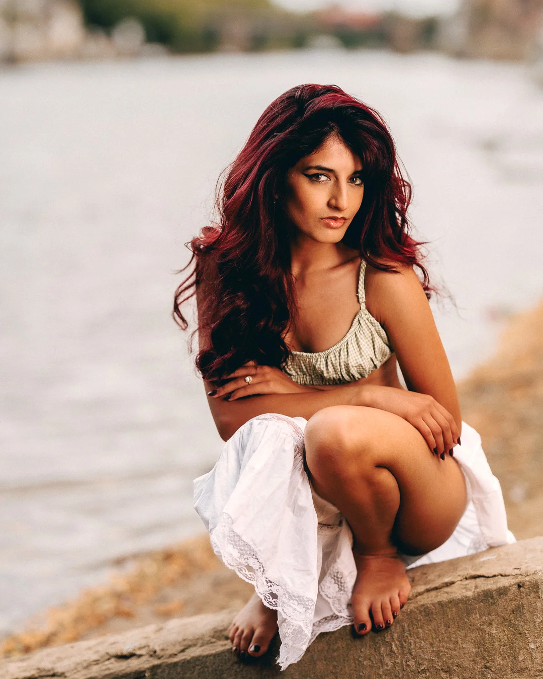 Woman with long red hair posing in a white skirt and light top by a riverbank.
