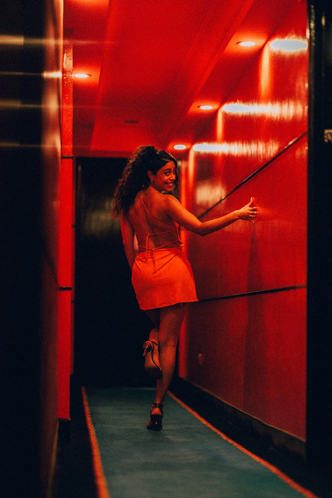 A woman in a short orange dress posing in a red-lit hallway, looking back over her shoulder.