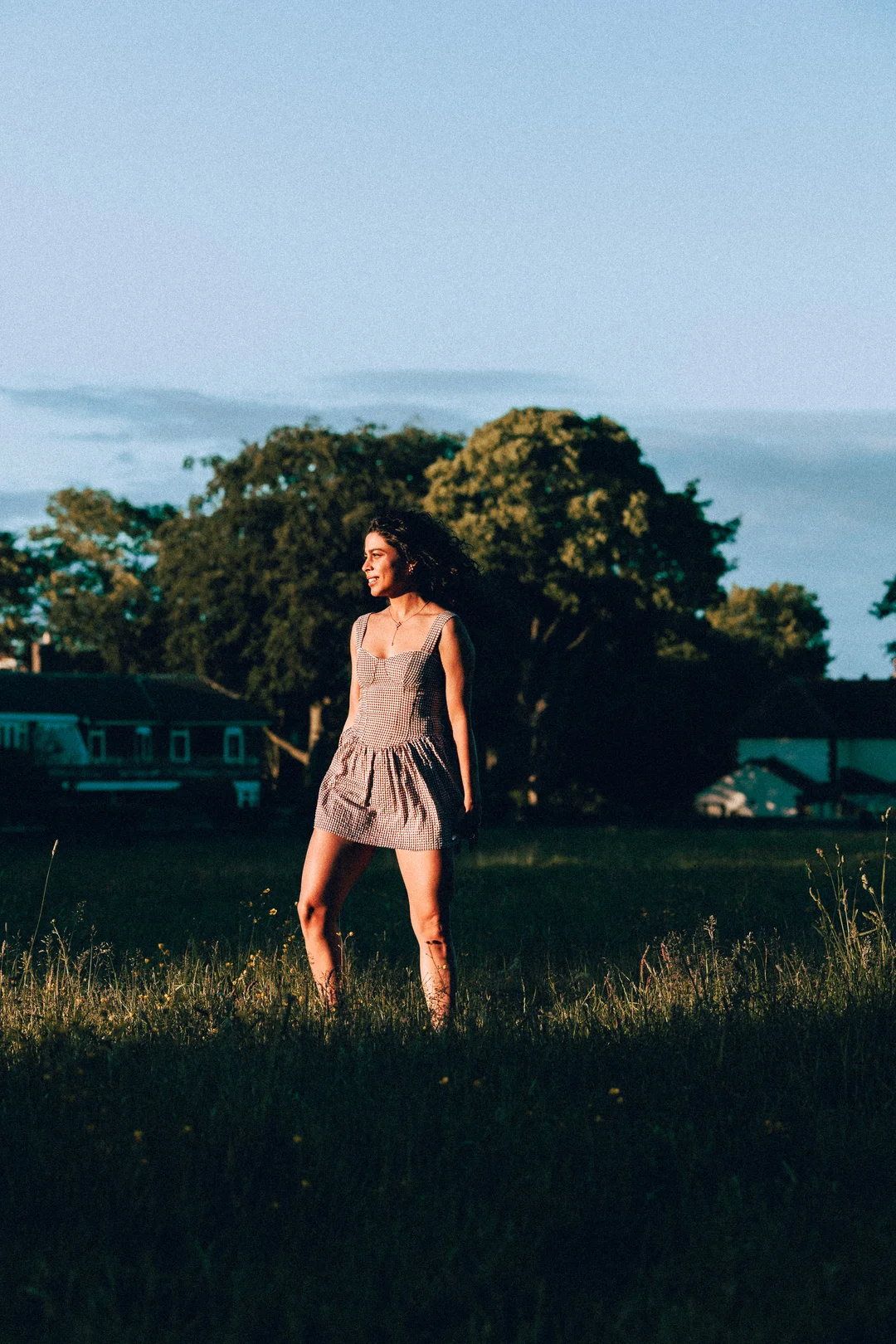 Woman in a red and white gingham dress standing in a sunlit grassy field.