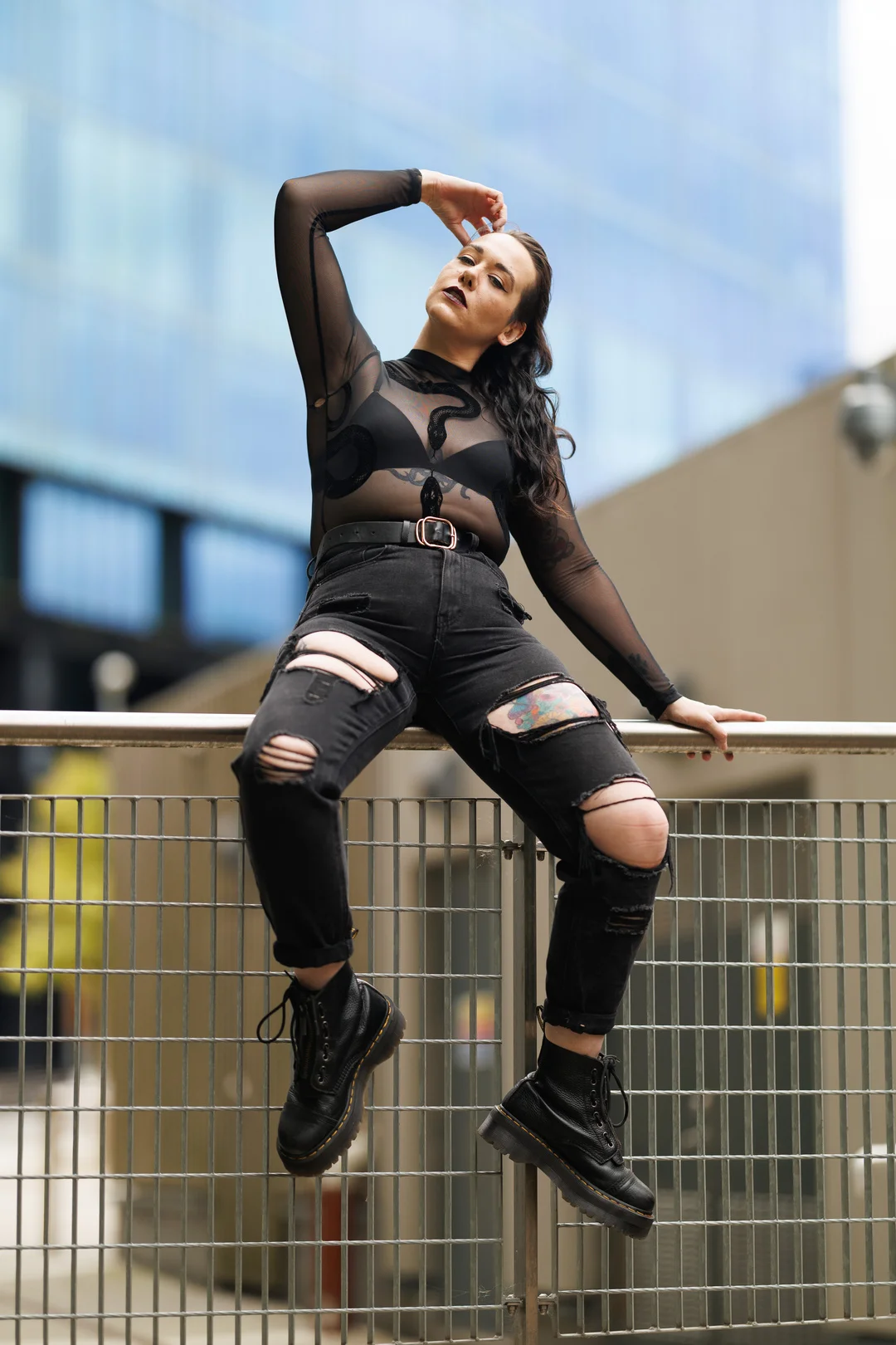 Woman in a black sheer top, ripped black jeans, and boots posing on an urban railing.