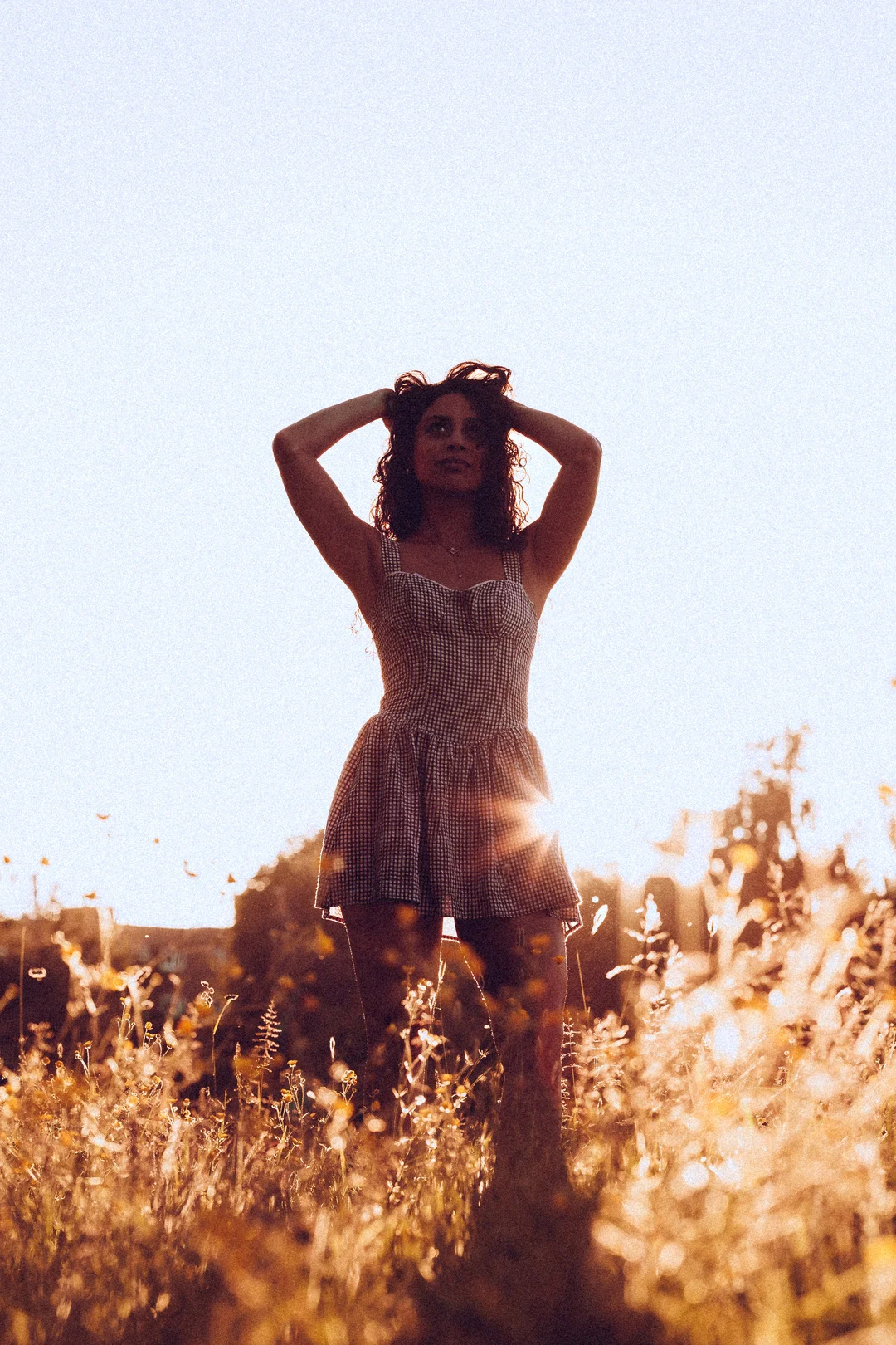A woman in a checkered dress standing in a sunlit field of wildflowers, hands in her hair during golden hour.