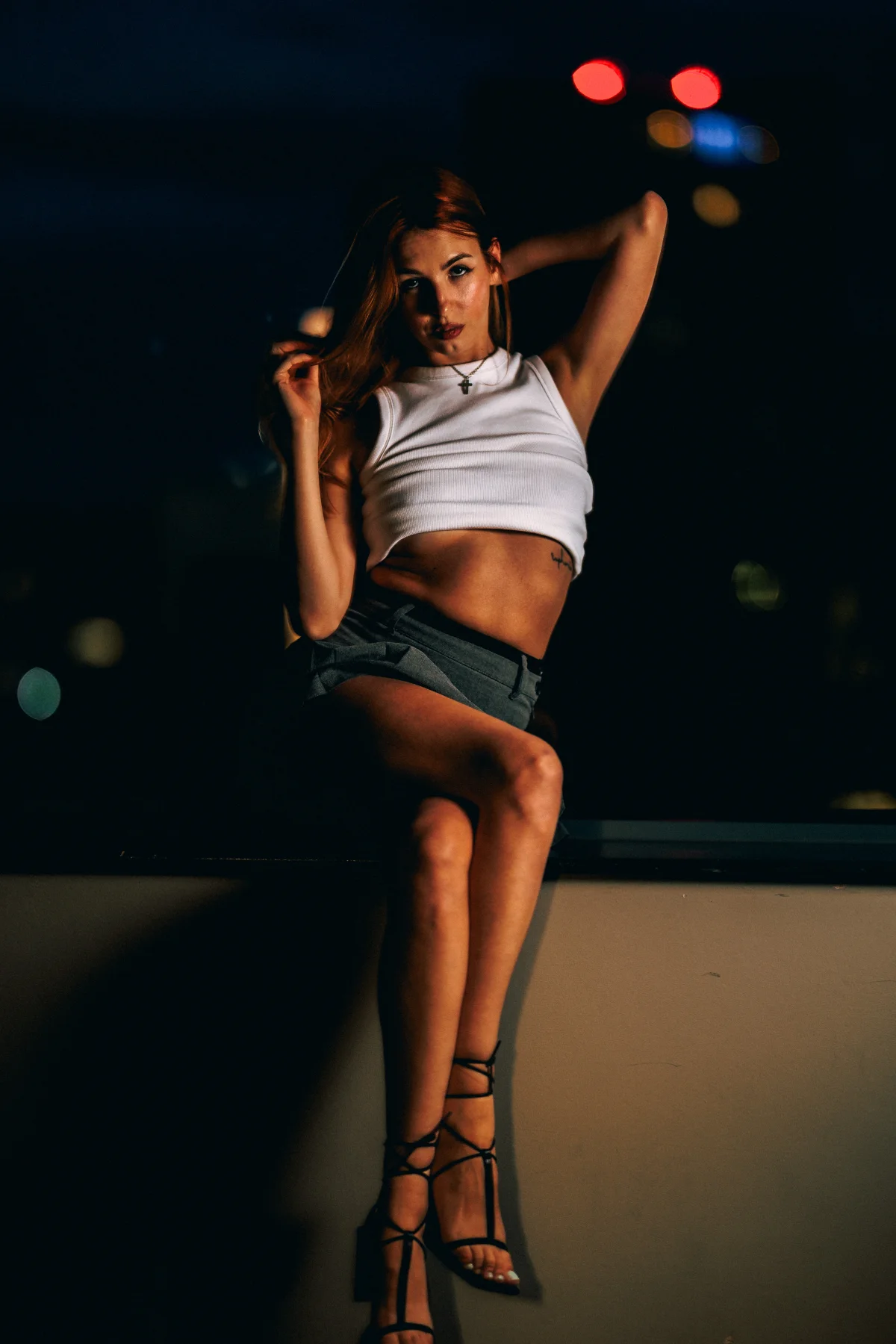 A woman in a white crop top and grey mini skirt posing on a ledge at night with blurred city lights behind her.