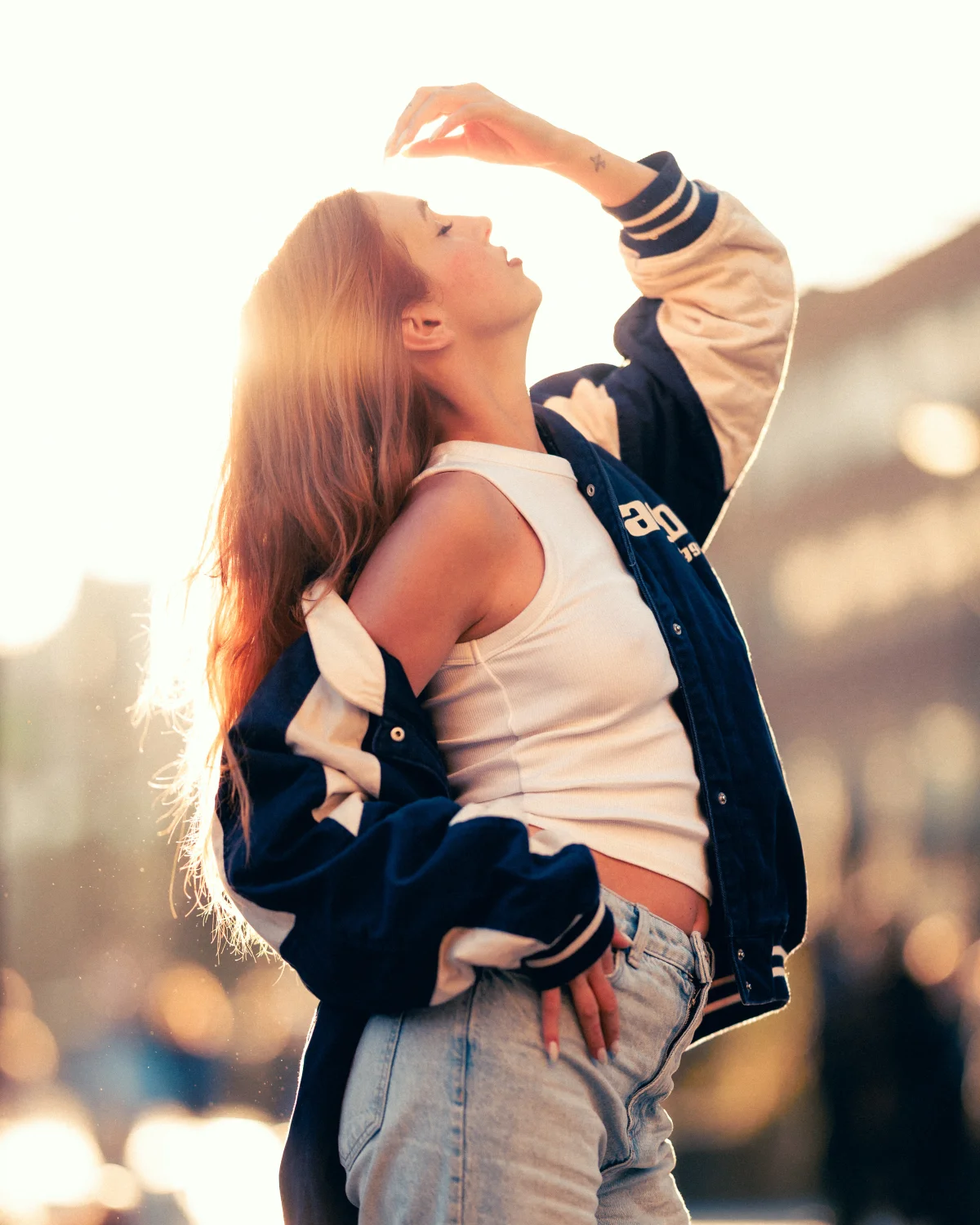 A young woman with long reddish-brown hair posing in a blue and white varsity jacket at sunset.
