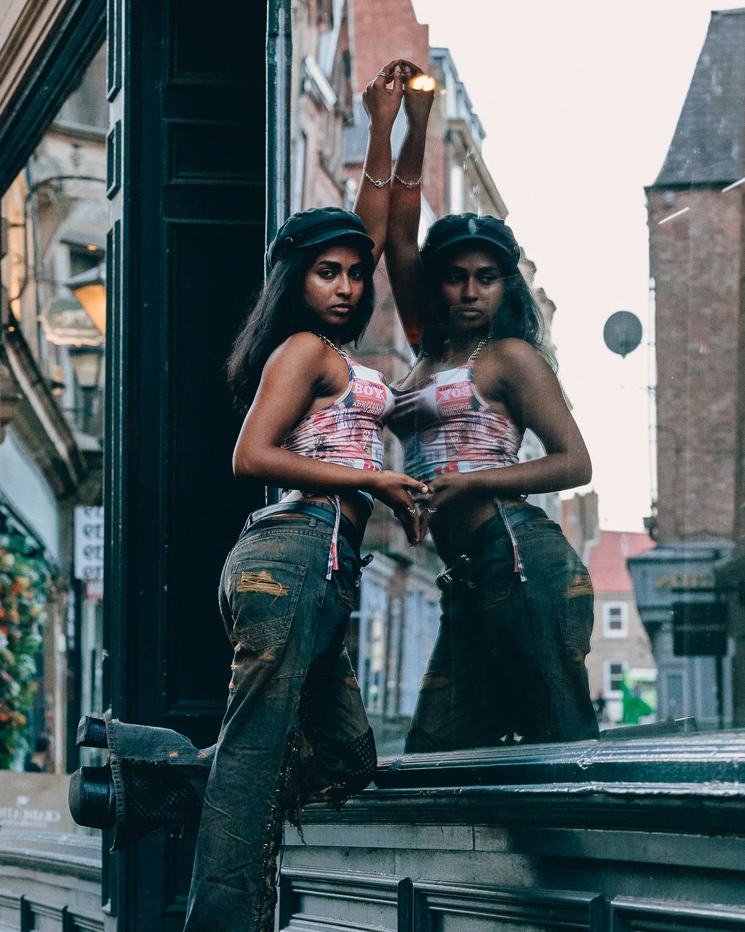 A woman in a crop top and distressed jeans posing with her reflection in an urban window.