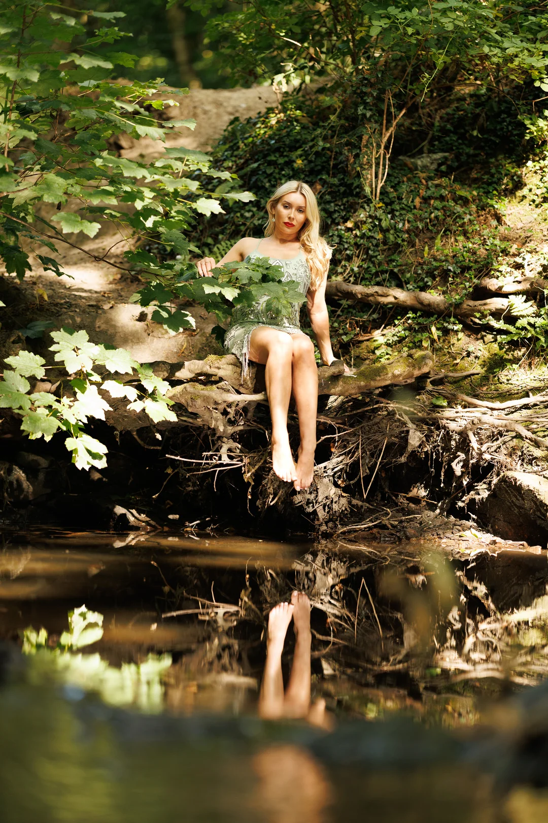 Blonde woman in a shimmering green dress sitting on a log by a stream in a lush forest, with a reflection in the water.