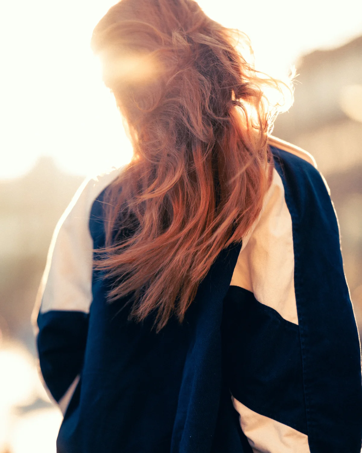 Back view of a person with long reddish-brown hair in a blue and white jacket, backlit by bright sunlight.