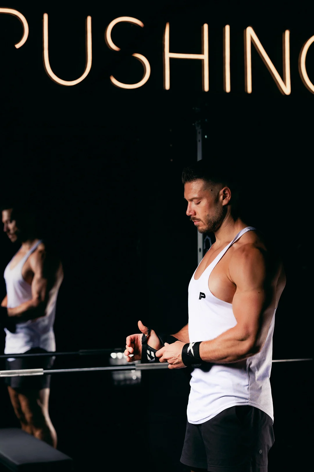 Muscular man in white athletic wear adjusting wrist wraps in a dark gym under a neon 'PUSHING' sign.