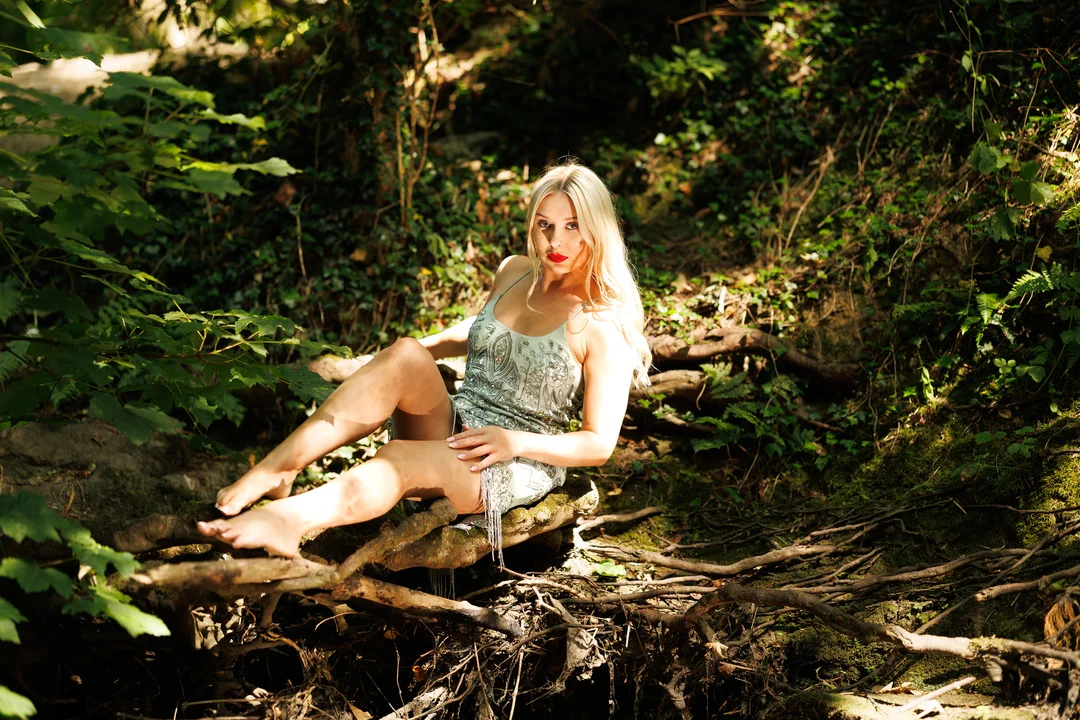 A blonde woman in a silver sequined dress posing on a fallen log in a lush green forest.