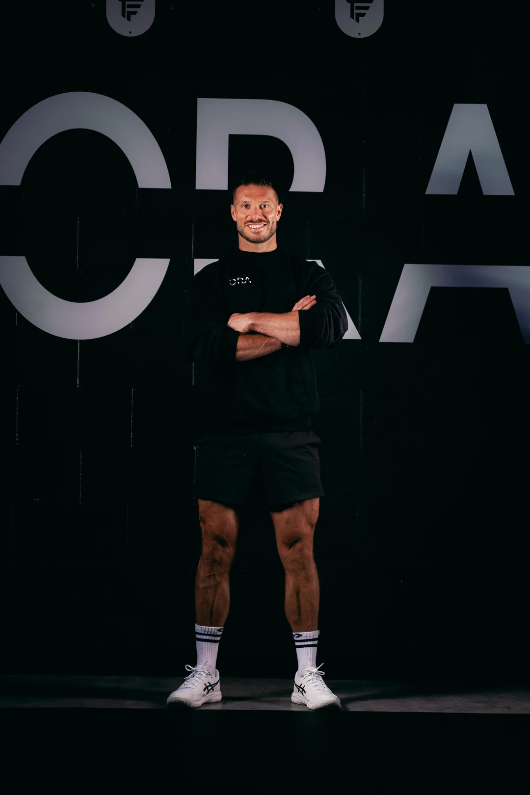 Full-body portrait of a muscular man in black athletic wear standing with arms crossed against a black branded wall.
