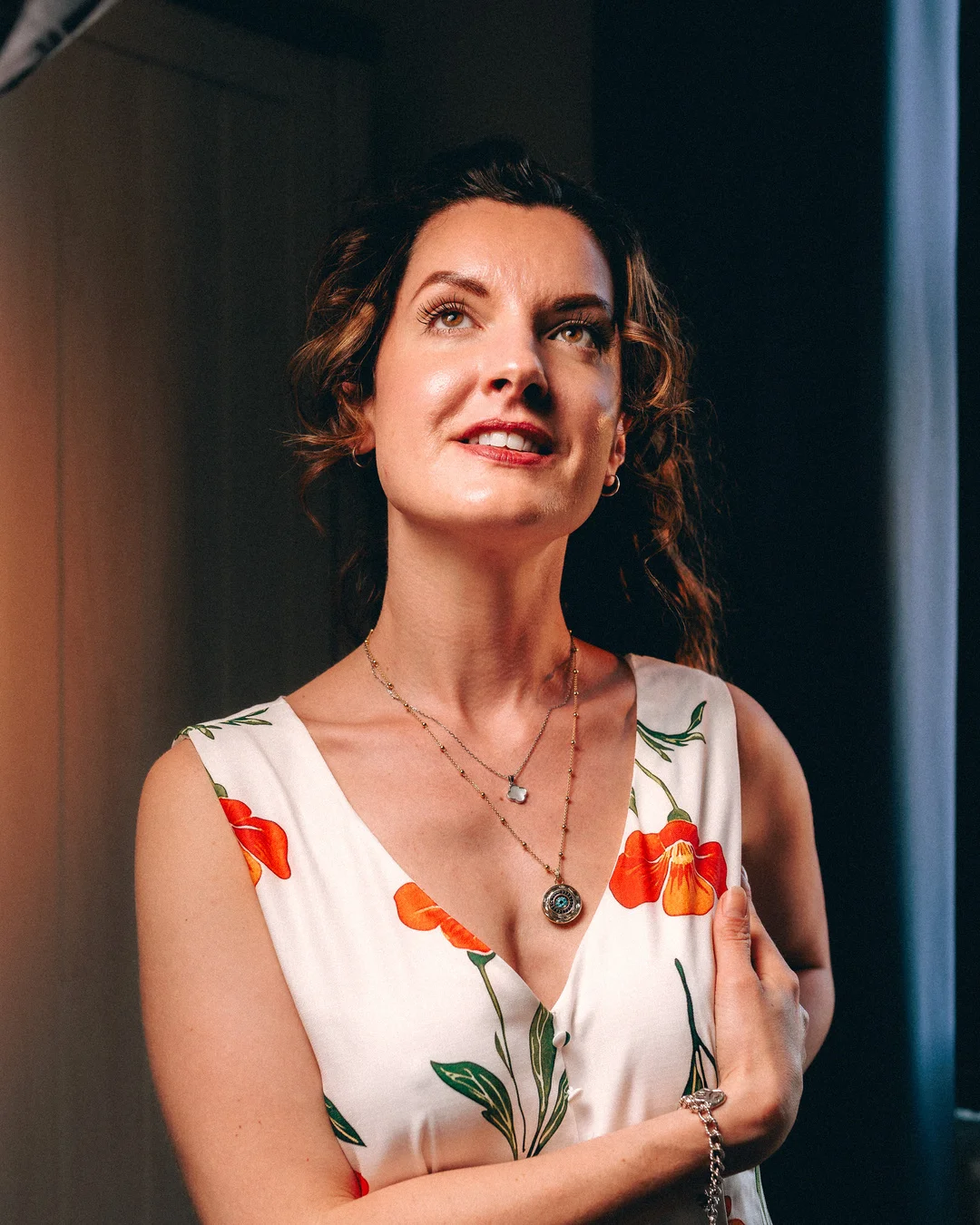 Woman with curly brown hair in a white floral dress looking upwards with a soft smile.