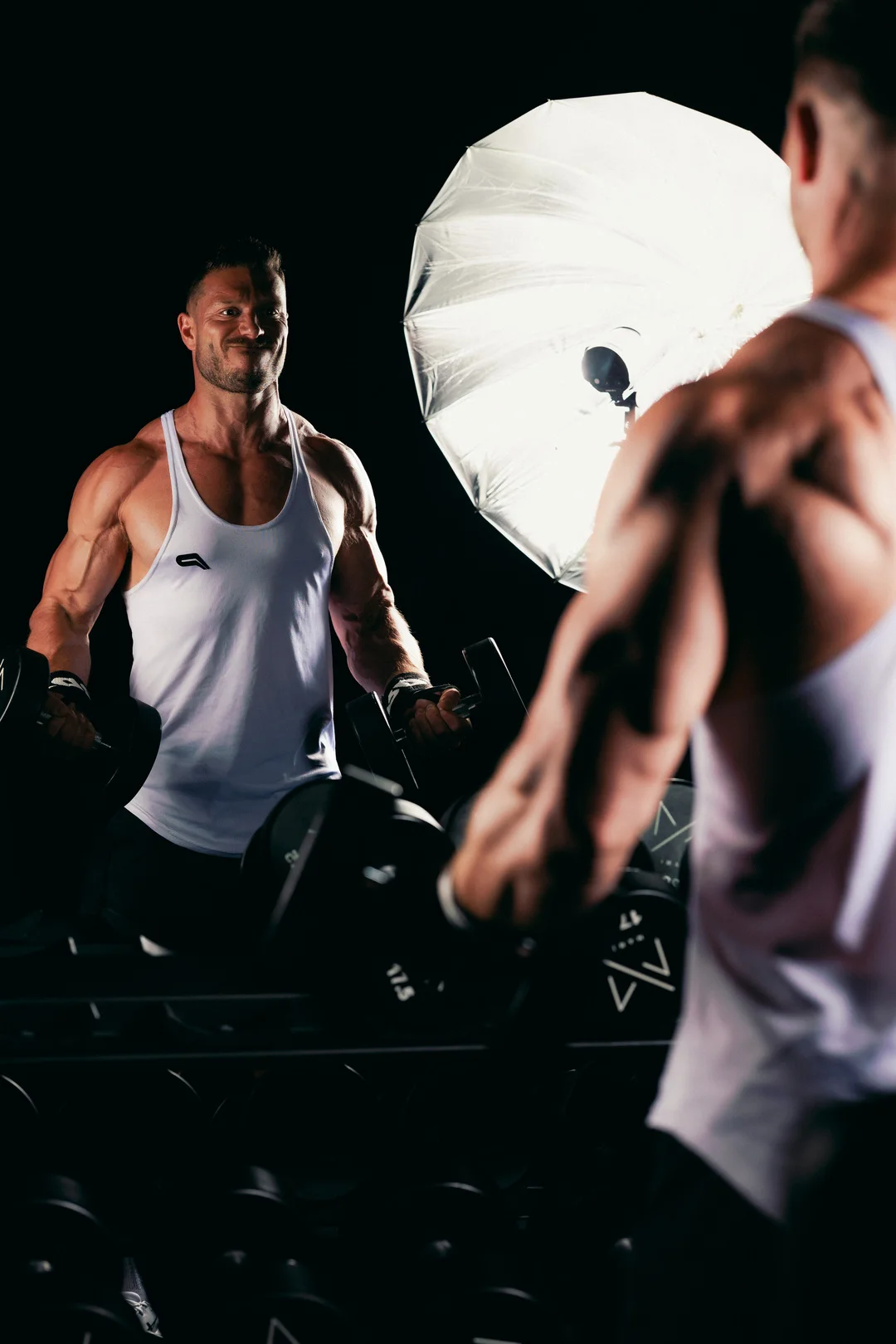 Muscular man in a white tank top posing with dumbbells in a dark gym, reflected in a mirror.