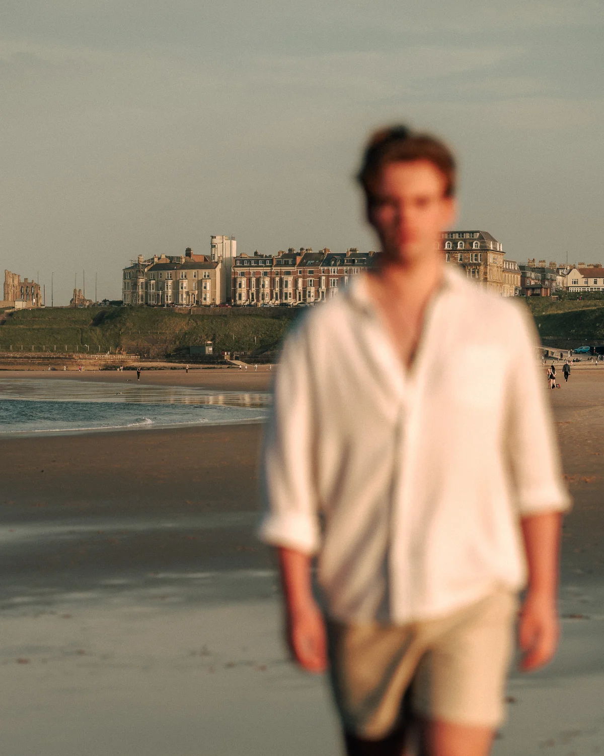 A blurry, artistic portrait of a man in a white shirt on a beach with coastal buildings in the background.