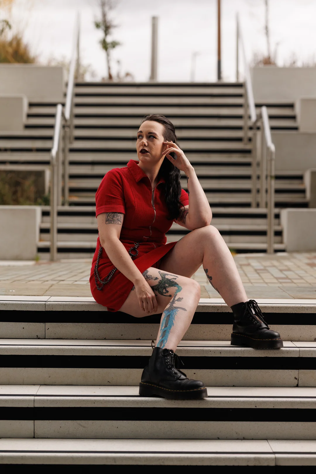 Woman with tattoos in a red dress and black boots sitting on outdoor concrete stairs.