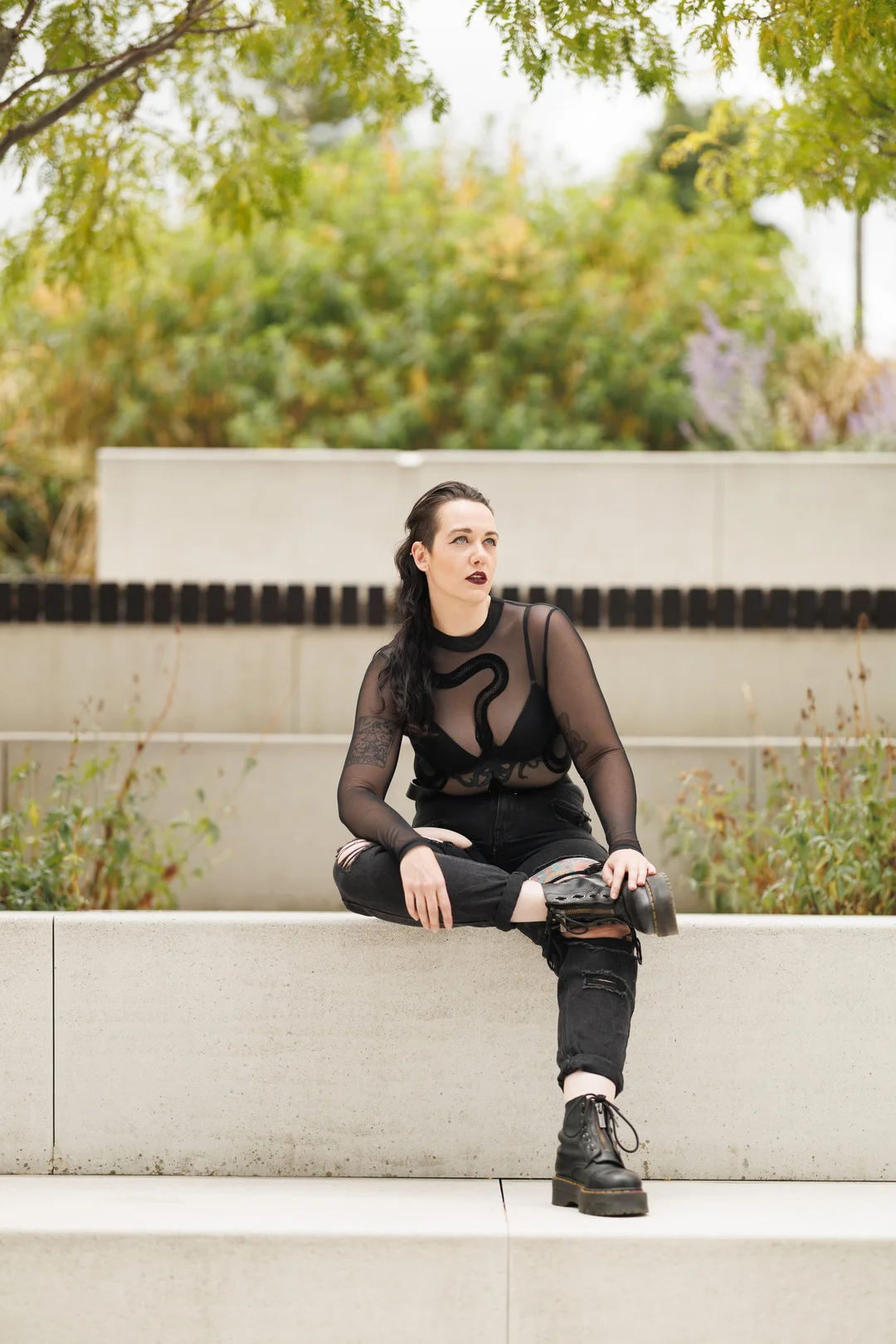 A person with dark makeup and hair wearing a black mesh top and ripped jeans sitting on a concrete ledge.