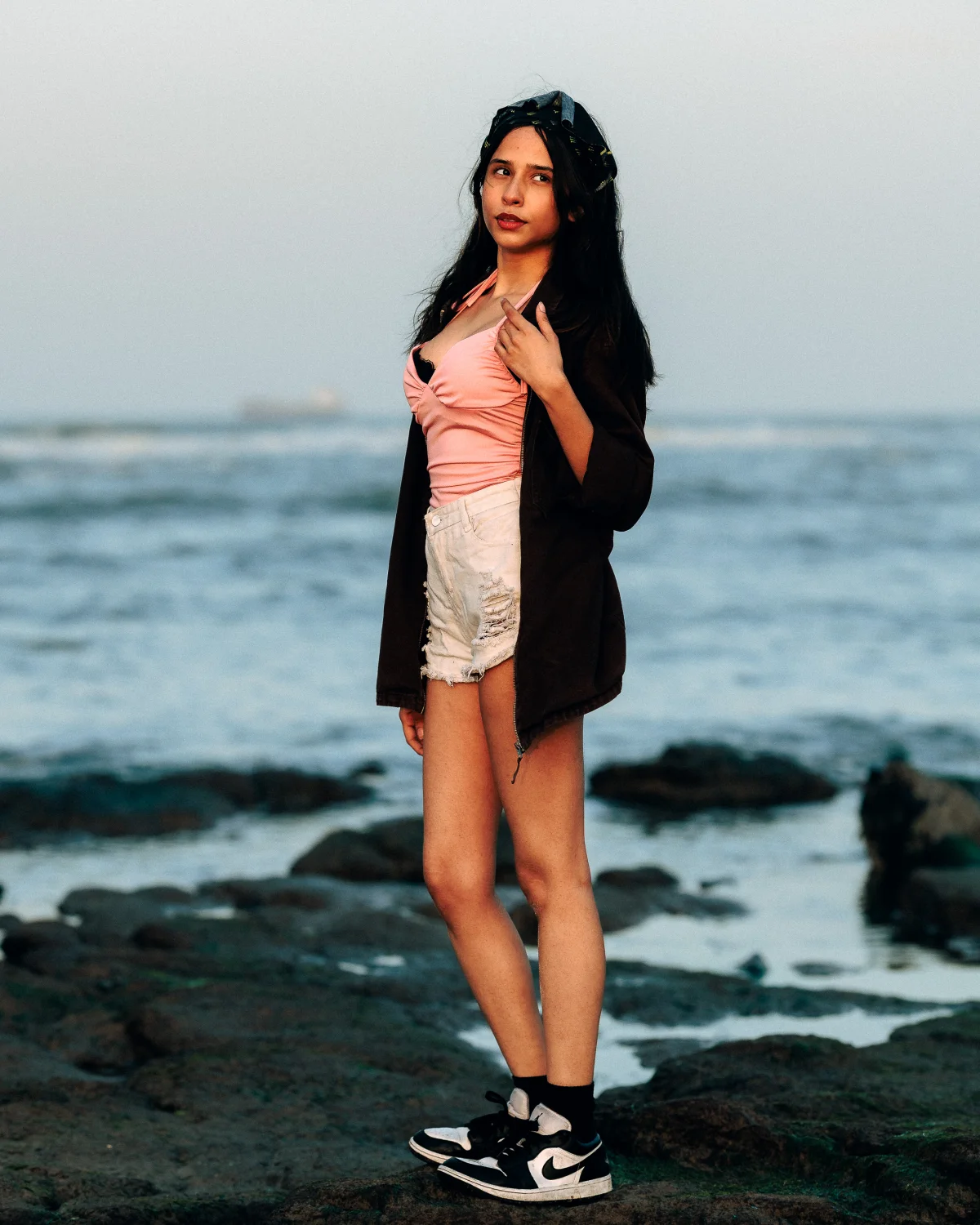 Full-body fashion portrait of a young woman posing on a rocky beach at dusk.