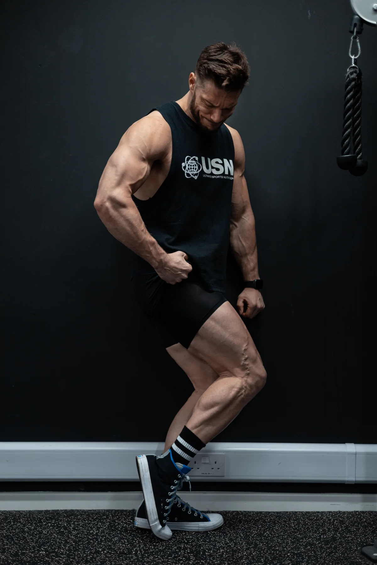 Muscular man in black fitness gear posing against a dark wall in a gym.