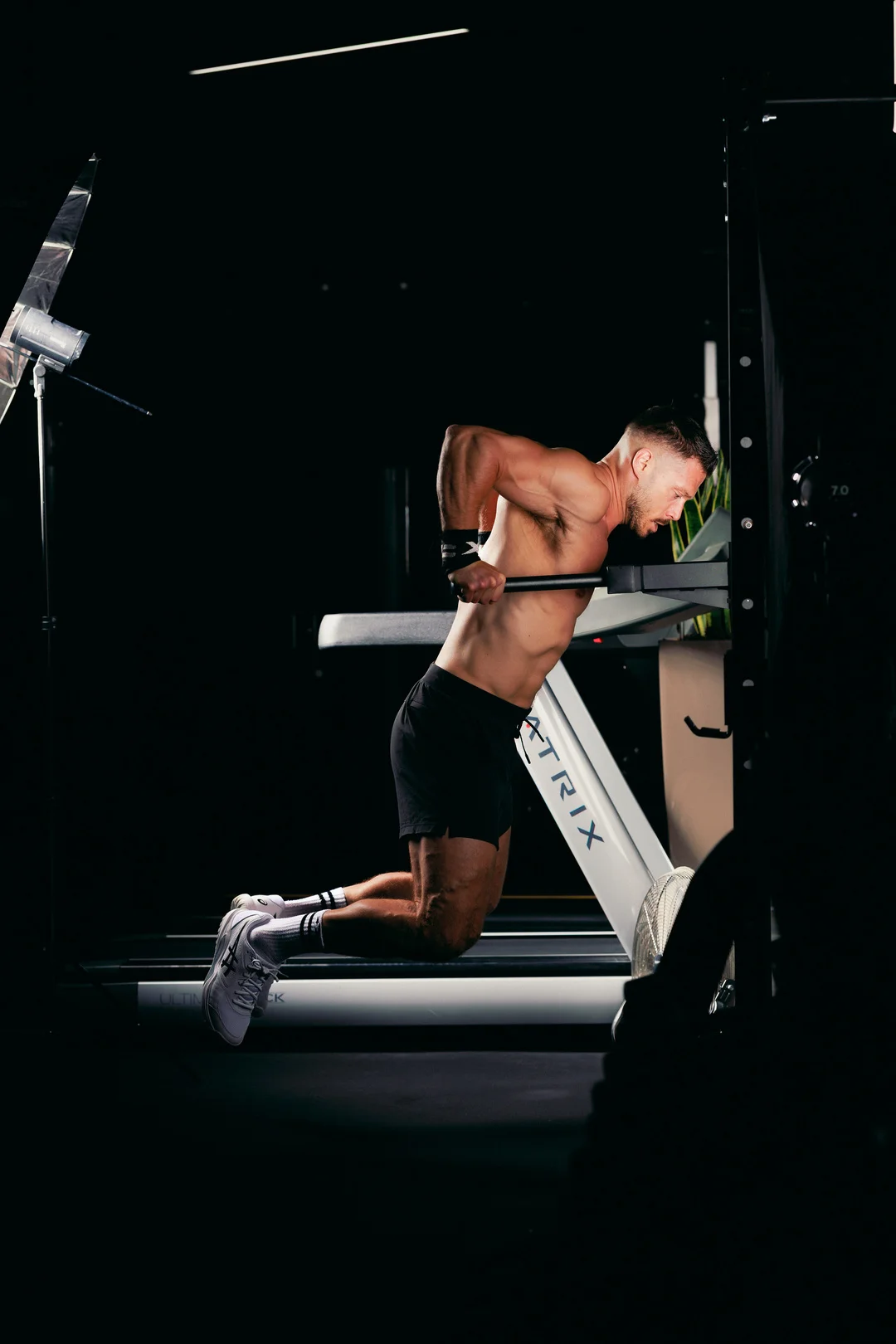 A shirtless athletic man performing dips on a gym machine against a dark, moody background.