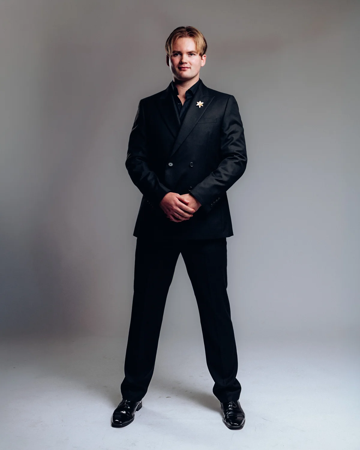 Full-body studio portrait of a man in a black double-breasted suit against a neutral grey background.