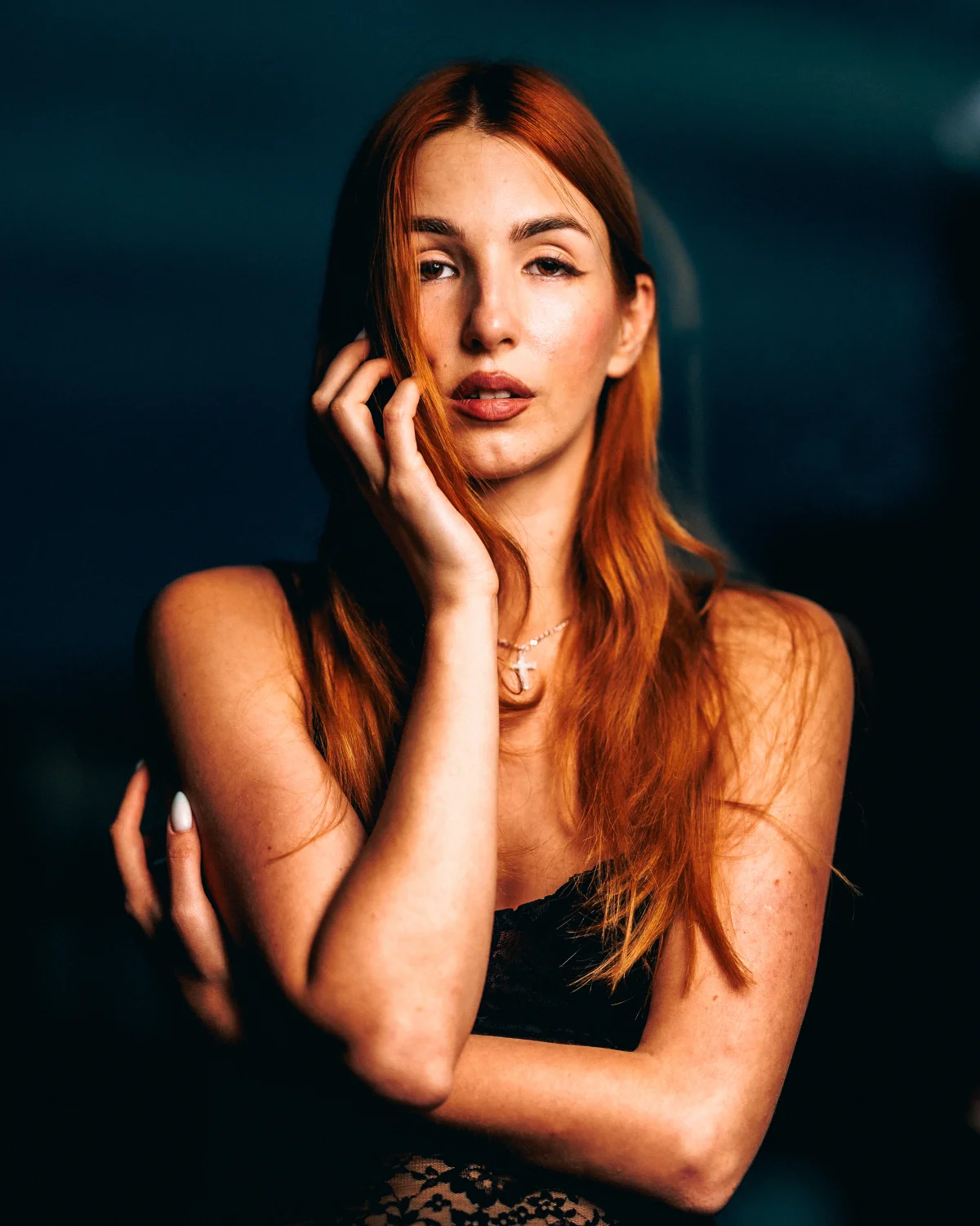 Moody portrait of a woman with long red hair against a dark background, hand touching her face.