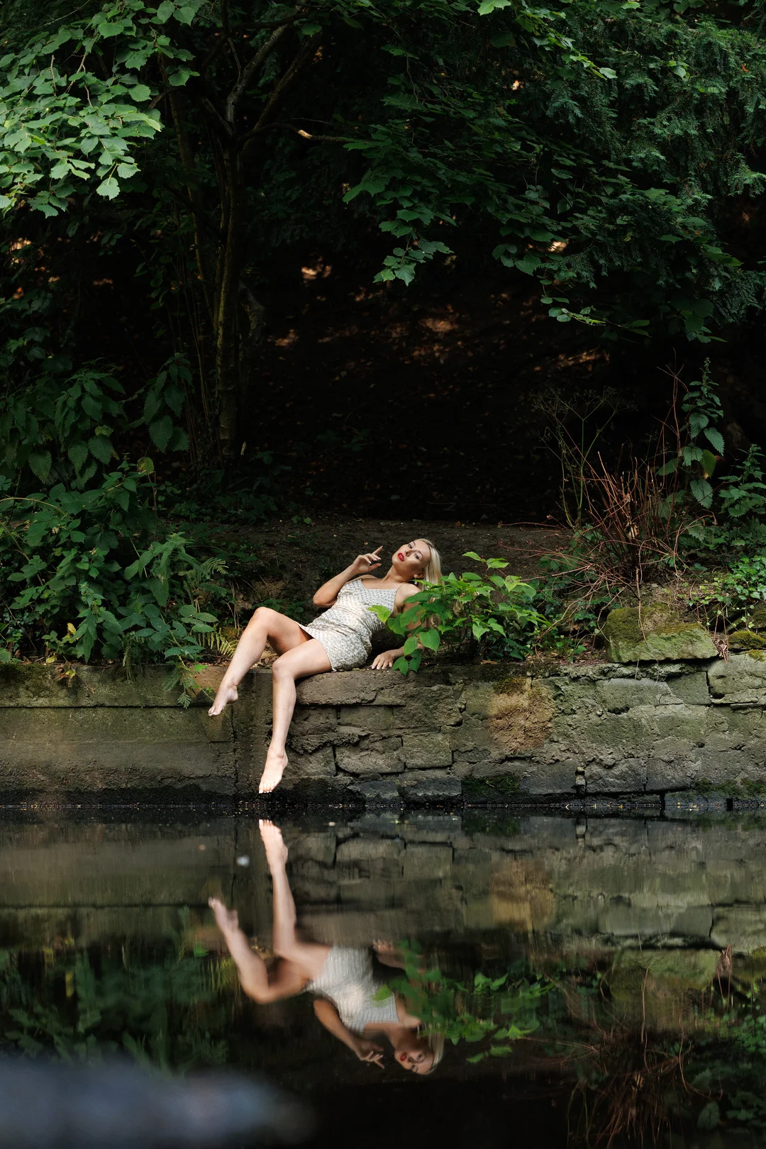 Blonde woman in a short gold sequin dress posing on a stone wall by a reflecting pond in a forest.