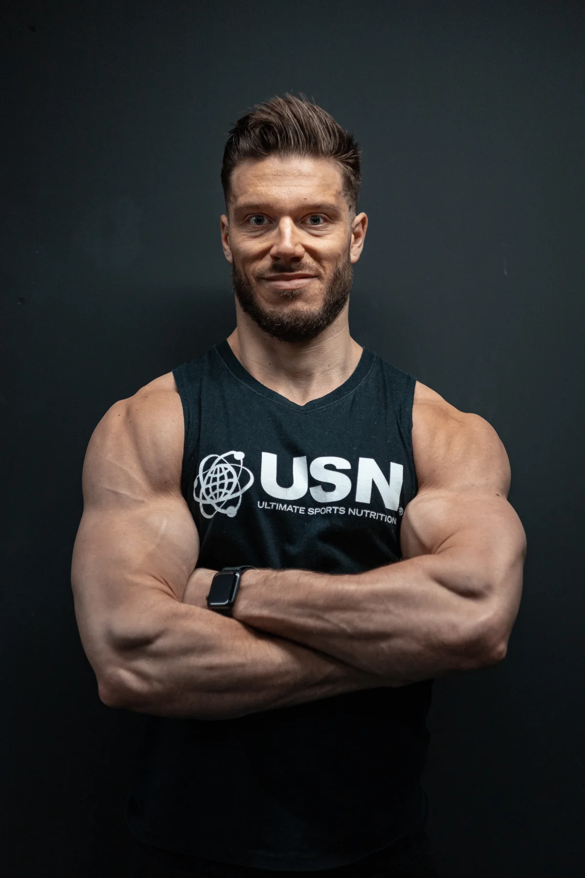 Muscular man with a beard wearing a black USN tank top, arms crossed, against a dark studio background.