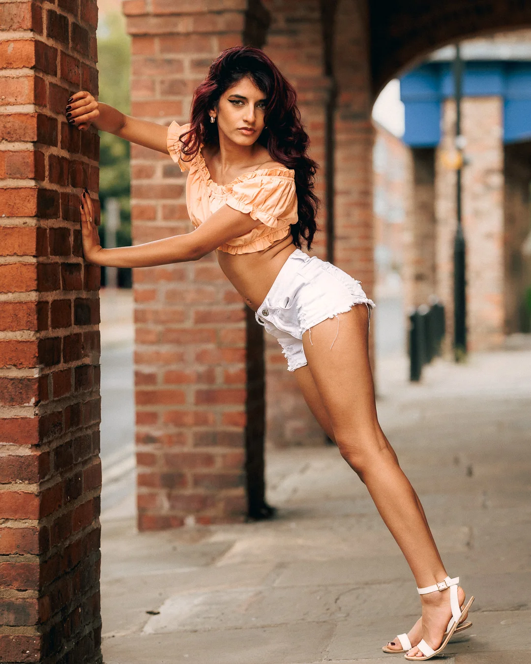 Woman with red hair posing against a brick wall in an urban setting, wearing a peach crop top and white denim shorts.