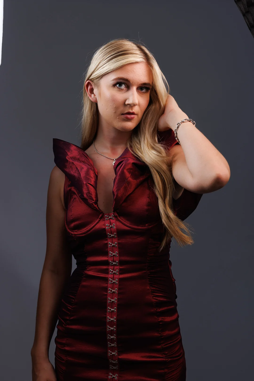 Blonde woman in a deep red satin dress posing against a grey studio background.