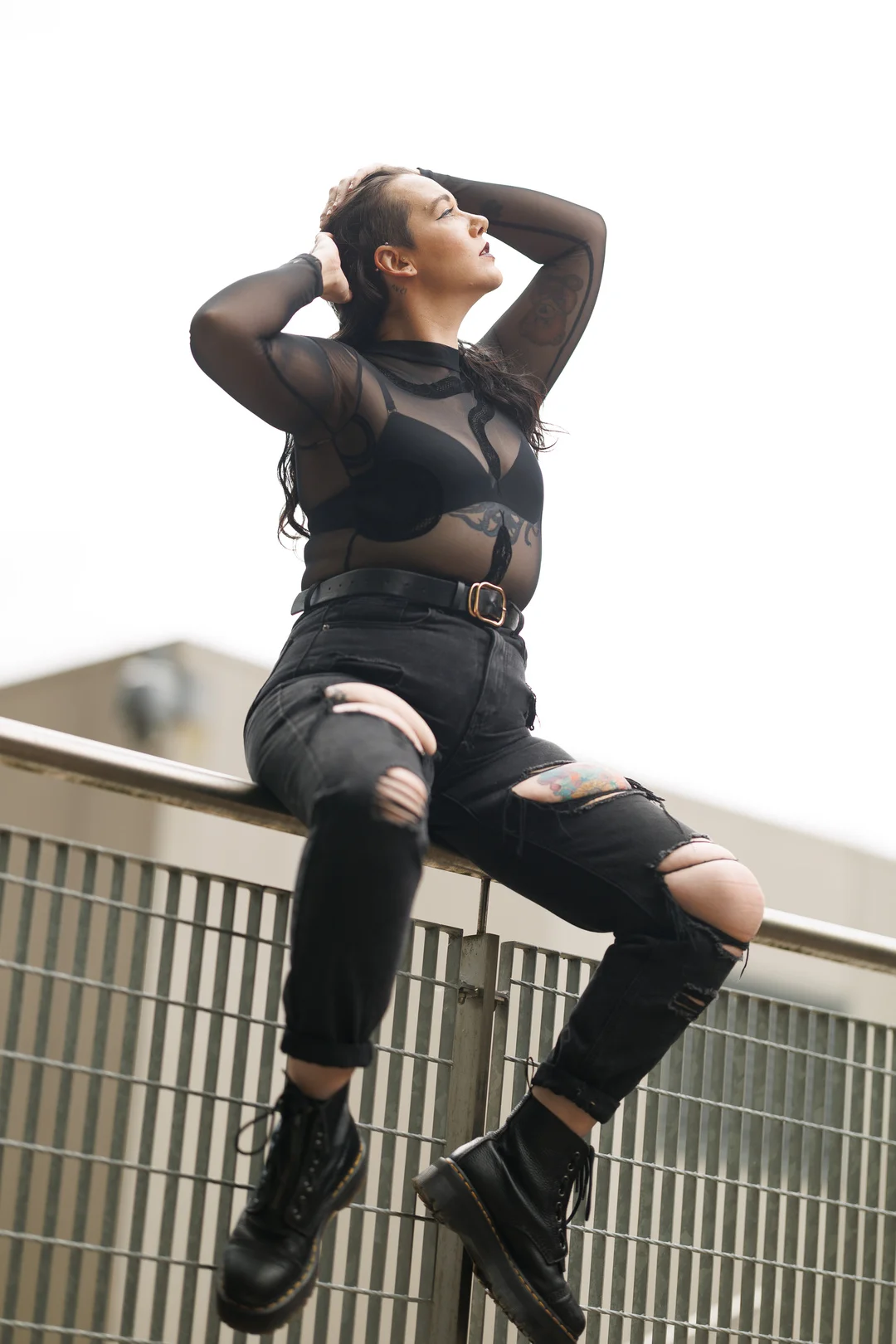 A tattooed person in a sheer black top and ripped black jeans posing on a metal railing against a bright sky.