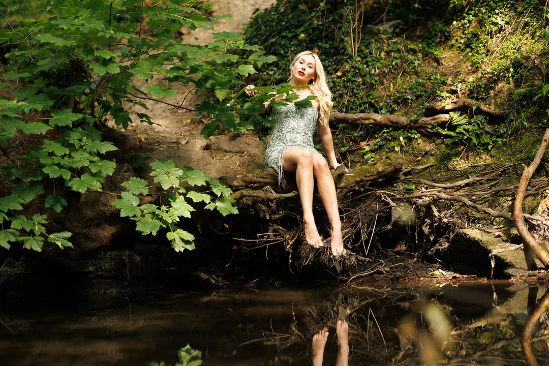 A blonde woman in a silver sequined dress sitting on a log by a stream in a lush forest.